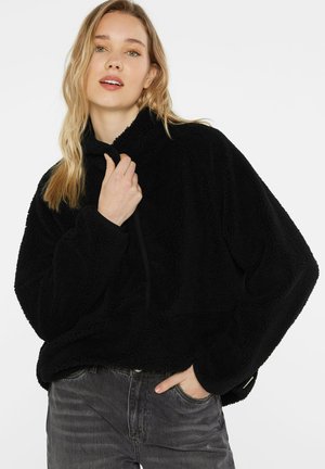 Blonde woman wearing a black fleece pullover with quarter zip and high collar, paired with dark gray jeans, standing against a white background.
