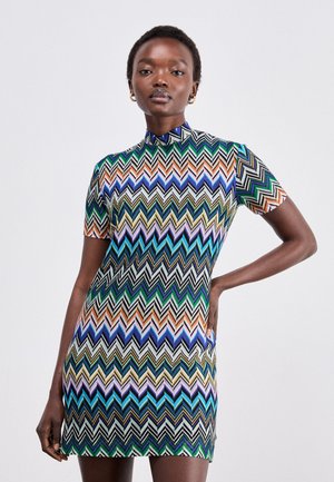 Short-sleeve mini dress featuring a multicolored zigzag pattern in greens, blues, and oranges, with a high collar and fitted design.