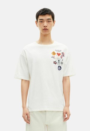 Camiseta branca oversized em algodão, com vários apliques coloridos no lado esquerdo do peito, mangas curtas e decote redondo.