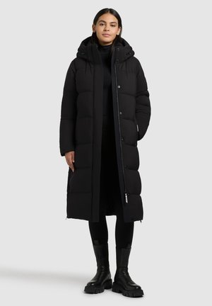 Schwarzer langer Steppmantel mit Kapuze, der ein gestepptes Design, vordere Druckknöpfe, seitliche Reißverschlüsse und eine gerade Silhouette aufweist. Kombiniert mit schwarzen Leggings und Stiefeln.