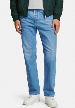 Man wearing light blue straight-leg jeans, white sneakers, white shirt, and dark green zip-up jacket, standing against white background.