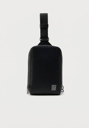 Μαύρο δερμάτινο τσαντάκι με φερμουάρ, με λεία υφή, ορθογώνιο σχήμα, ανθεκτικό ιμάντα και μεταλλικό λογότυπο στη μπροστινή πλευρά.