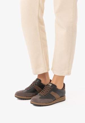 Personne portant un pantalon beige et des baskets décontractées marron et noires à lacets, debout sur une surface blanche.