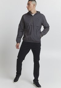 Gray, lightweight pullover jacket with a hood, half zipper, elastic cuffs, and hem. Paired with black pants and black sneakers.
