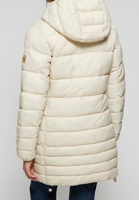 Doudoune couleur crème avec des sections matelassées horizontales, capuche et petite étiquette logo ; comporte un cordon de serrage à l'ourlet et une texture lisse.