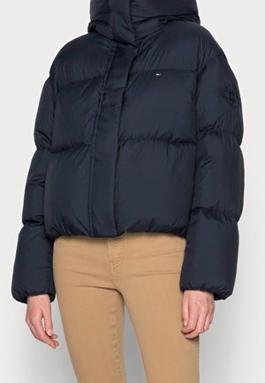 Osoba ubrana w granatową pikowaną kurtkę z wysokim kołnierzem i beżowe spodnie, stojąca na tle jasnego, jednolitego tła.