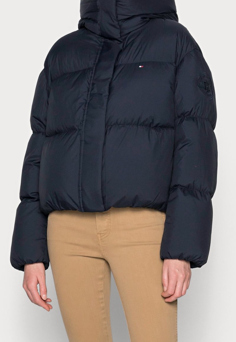 Persona usando una chaqueta acolchada azul marino con cuello alto y pantalones beige, de pie contra un fondo claro y liso.