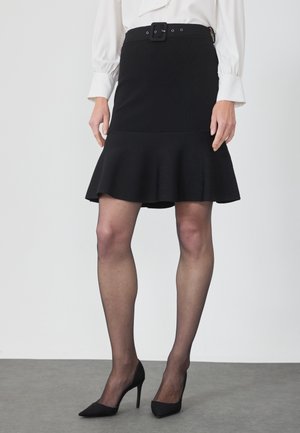 Vrouw met een zwarte uitlopende rok met riem, doorzichtige zwarte panty's, zwarte hoge hakken en een witte blouse met omgeslagen mouwen.