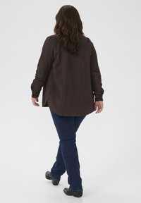 Brun mockajacka med lång ärm, lätt bakåtböjning, kombinerad med mörkblå jeans och svarta skor. Enkel och mjuk textilstruktur.