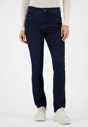 Person trägt dunkelblaue Jeans mit geradem Bein, ein helles Langarmshirt und weiße Sneakers und steht vor einem schlichten Hintergrund.