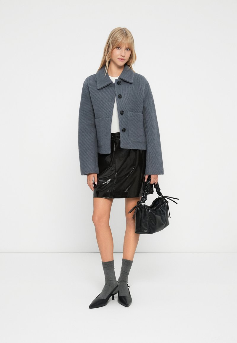 Junge Frau in grauem knöpfbarem Jackett, schwarzem glänzenden Rock, grauen Socken und schwarzen Schuhen mit Absatz, die eine schwarze Handtasche vor einem weißen Hintergrund hält.