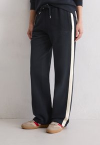 Person, der schwarze weit geschnittene Hosen mit weißen Seitenstreifen und beigefarbene Sneakers mit roten Akzenten trägt und auf einem hellen Boden steht.