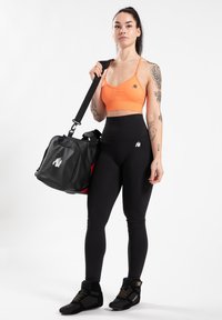 Zwarte en rode sporttas met schouderband, gecombineerd met een oranje sportbeha en zwarte hoge leggings, tentoongesteld op een model.