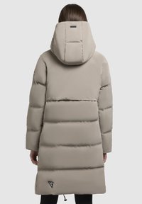 Gaisa pūkains mētelis gaiši bēšā krāsā ar lielu pildītu kapuci, horizontālu šuvju rakstu, elastīgu auklu apakšmalā un logotipu apakšējā kreisajā stūrī.