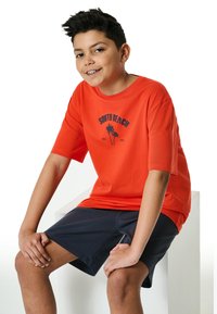 Red short-sleeve t-shirt with "SOUTH BEACH" graphic and palm tree design, paired with dark shorts; lightweight cotton material, relaxed fit.