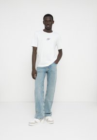 Valkoinen puuvillainen t-paita, jossa on pieni graafinen painatus rinnassa, yhdistetty vaaleansinisiin leveälahkeisiin farkkuihin ja valkoisiin lenkkareihin, joissa on harmaat yksityiskohdat.