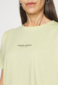 Světle žluté bavlněné tričko s kulatým výstřihem a krátkými rukávy. Na přední straně je natištěn text "CONTEMPORARY_SCANDINAVIAN."