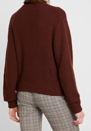 Person wearing a brown ribbed knit sweater and beige plaid pants, shown from the back against a plain light background.