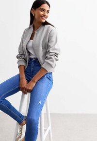 Chaqueta bomber gris claro con puños acanalados, usada sobre una camiseta blanca ajustada. Jeans azules de talle alto con detalles desgastados. Zapatillas neutras.