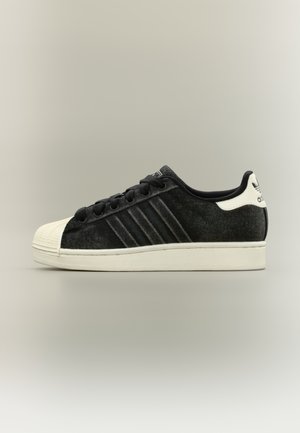 Czarny zamszowy sneaker z teksturowaną cholewką, białym gumowym noskiem, trzema paskami jako akcentami oraz płaskimi czarnymi sznurowadłami. Klasyczny niskoprofilowy fason.