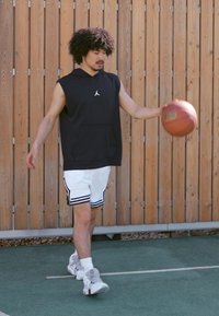 Sort ærmeløs hættetrøje med lomme, hvide shorts med sorte striber og grå sneakers. Spiller med en orange basketball på en bane.