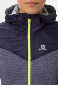 Marinblå lila träningsjacka med hög krage, med en klar gul dragkedja, grå strukturerad tyg och en logotyp på vänster bröst.