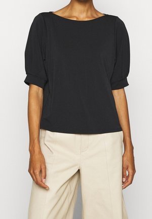 Vrouw draagt een zwarte korte mouwen blouse met omslagpofmouwen en beige hoge taille broek.