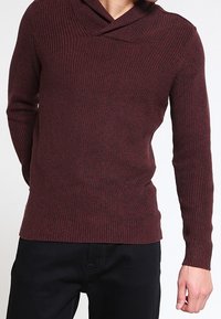 Pull en maille bordeaux avec col croisé, texture côtelée, manches longues et ourlet ajusté. Associé à un pantalon noir.