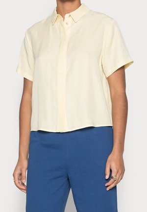 Chemise à col et manches courtes en tissu jaune doux. Présente une fermeture à boutons sur le devant et un ourlet court. Associée à un pantalon bleu.
