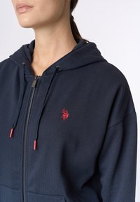 Felpa con zip di un blu navy realizzata in tessuto morbido. Presenta un piccolo logo rosso sul petto e corde di regolazione con accenti rossi.