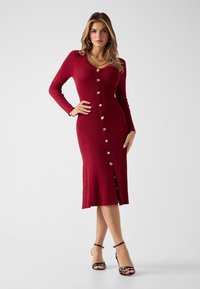 Strick-Midikleid in Burgunderrot mit gerippter Struktur, langen Ärmeln, V-Ausschnitt und goldenen Knöpfen vorne. Mit seitlichem Schlitz und figurbetonter Silhouette.