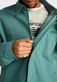 Giacca leggera color verde petrolio con cerniera frontale, colletto a punta e polsini con bottoni automatici. Presenta una tasca sul petto e un dettaglio sottile del logo. Indossata sopra una camicia grigia.