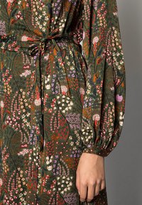 Groene jurk met een textuur en bloemmotieven in roze, wit en rood. Lange mouwen met volants aan de manchetten en een bijpassende strikriem in de taille.