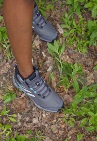 Scarpa da trekking grigia con tessuto testurizzato, sovrastrutture sintetiche, accenti blu e suola in gomma, posizionata su un terreno foglioso.