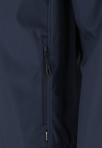 Marinblå jacka med en ficka med dragkedja och en texturerad dragkedja, slät tyg och en logotypmärke nära fållen.