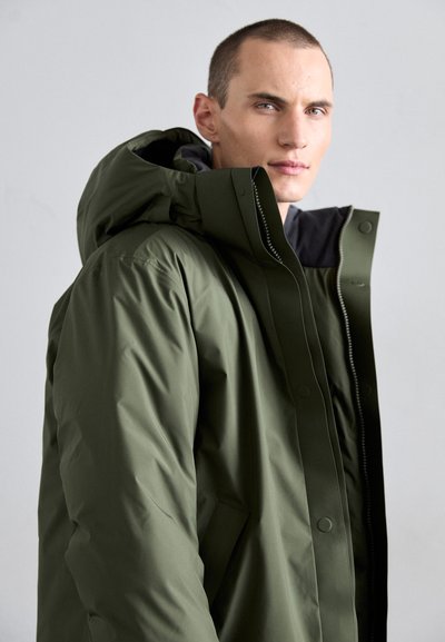 Olive green puffer jacket with a large hood, front zip, and snap closures. Features a textured exterior and a black inner lining.
