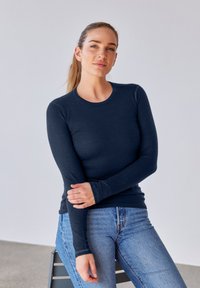 Marineblaue Langarmbluse in taillierter Form, hergestellt aus weichem Material; mit rundem Halsausschnitt und Ärmel in Handgelenklänge, kombiniert mit hellblauen Jeans.