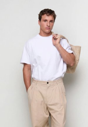 Young man wearing a white t-shirt and beige trousers with one hand in pocket, holding a beige bag over his shoulder against a plain background.
