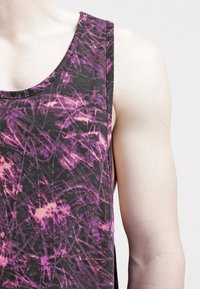 Černý tílko s abstraktním vzorem v purpurové a růžové barvě, měkká látka a široké výstřihy na pažích. Detailní pohled na rameno a horní část paže.