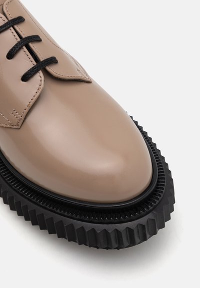Beige leather shoe featuring a glossy finish, black rubber sole with a chunky, ridged design, and classic lace-up closure with black laces.
