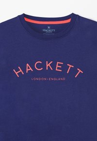 Námořnická modrá bavlněná tričko s kulatým výstřihem. Na přední straně se nachází výrazné korálové logo "HACKETT" a text "LONDON • ENGLAND" pod ním v zakřiveném uspořádání.