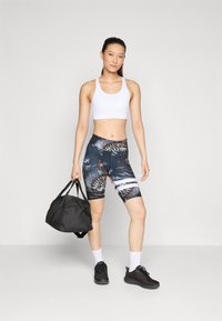 Čierna taška na cesty, biele športové podprsenky, vzorované legíny s vysokým pásom s tmavým základom a farebnými akcentmi. Čierne športové topánky a biele ponožky.