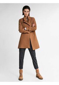 Abrigo de lana marrón con botones de doble broche, combinado con pantalones de cuadros y botines de tobillo color marrón claro. El abrigo tiene un diseño estructurado y bolsillos con solapa.