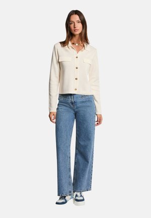 Veste crème à boutons avec deux poches avant, associée à un jean en denim large bleu clair et des baskets bleu et blanc.