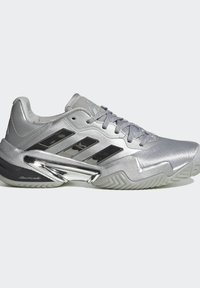 Strieborná Adidas nízka teniska s čiernymi pruhmi, sivými šnúrkami a textúrovanou podrážkou, s chrómovým detailom blízko päty.