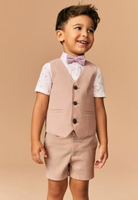 Costume trois pièces rose clair composé d'un gilet, d'une chemise à motif floral et d'un short. Comprend un nœud papillon et des boutons sur le gilet.