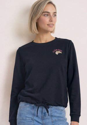 Femme aux cheveux blonds courts portant un sweat bleu marine avec "Le Café Paris" et un croissant brodé sur la poitrine, associé à un jean bleu clair.