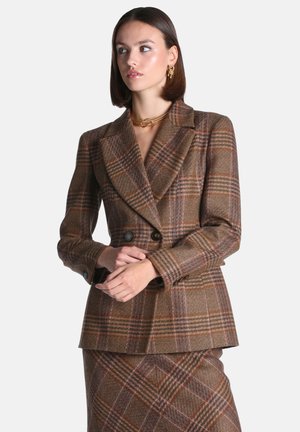 Femme portant un blazer croisé marron à carreaux et une jupe assortie, avec des boucles d’oreilles créoles en or et un collier en or torsadé, regardant sur le côté.