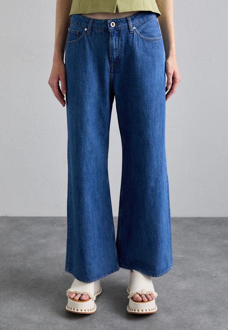 Jeans de denim de pierna ancha en un lavado azul medio, con una cintura alta, cinco bolsillos y un ligero desvanecimiento. Combinados con sandalias de plataforma en color crema.