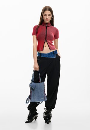 Mladá žena stojí před bílým pozadím, oblečená v červeném zkráceném topu na zip, černých kalhotách s modrým pasem, černých volných botách a drží džínovou tašku na rameno.
